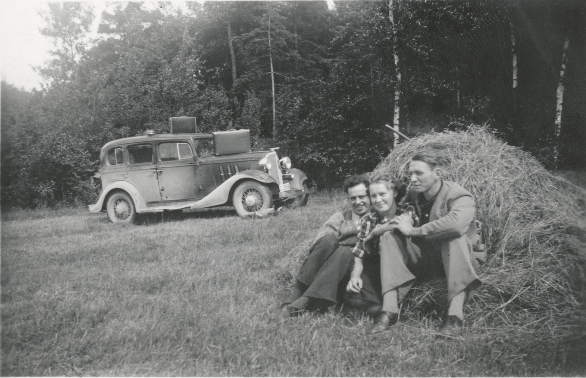 To unge menn og en dame på biltur mot Sverige, tar en rast i bakken ved høysåte. Chevrolet 1933-modell. Eier av bilen i 1935 var Magnus Bråthe.
