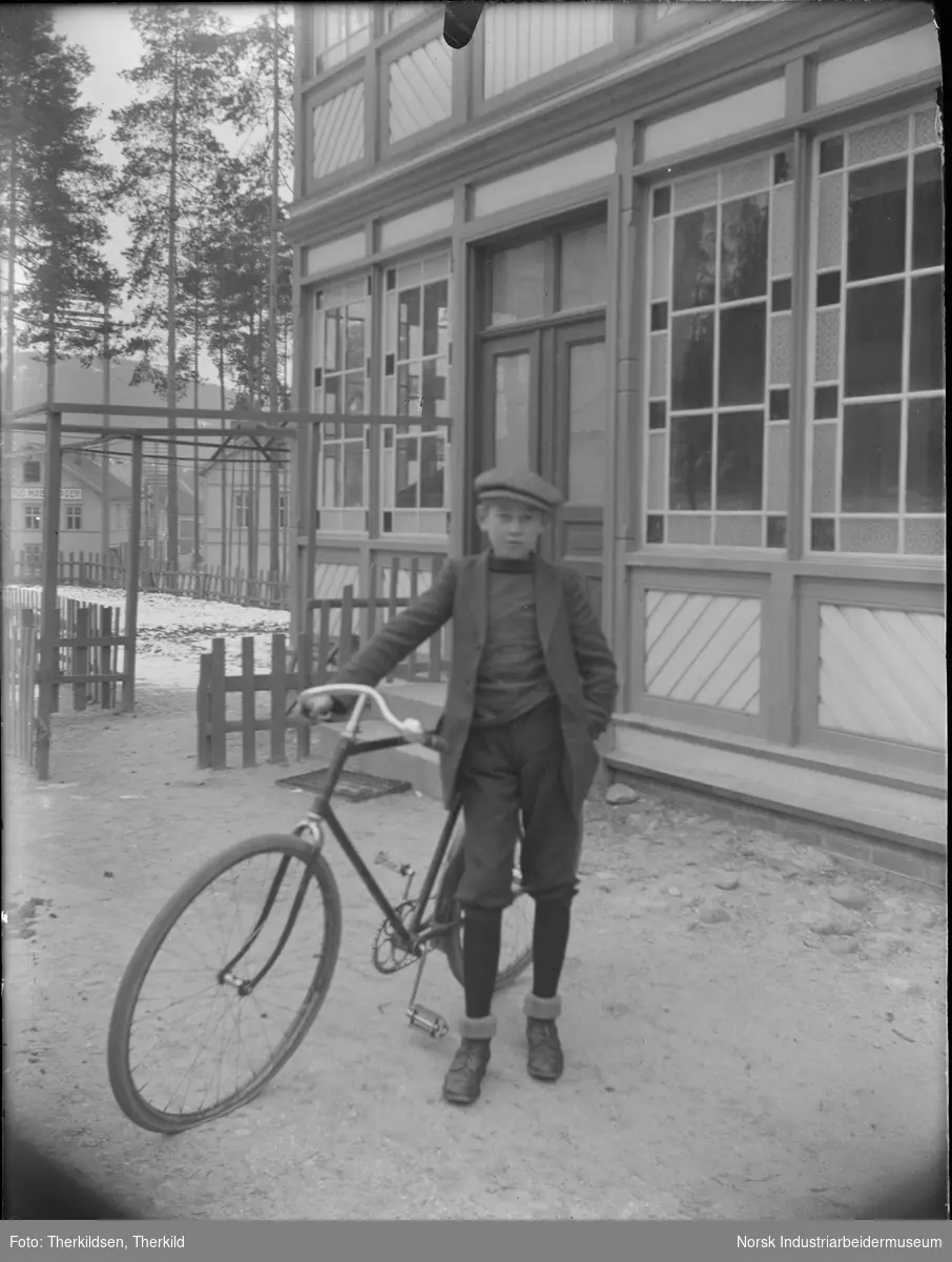 Gutt med sykkel kledd i knickers og sixpence stående foran Solvang, senere kalt Hellandgården. Bygningen har dobbel inngangsdør og smårutete vinduer i deler av fasaden. Bygg for møbellager i bakgrunnen.