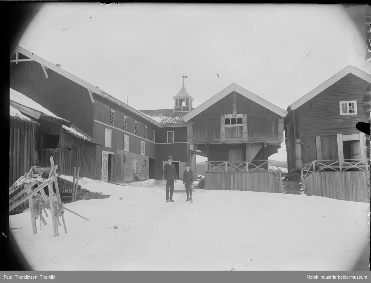 Mann og gutt stående på gårdstun på Søndre Landsverk. Fjøsbygning med matklokke, loft med utvendig trapp og 2 etasjes bur med innkledd svalgang. Hesteslede stående i snøen.
