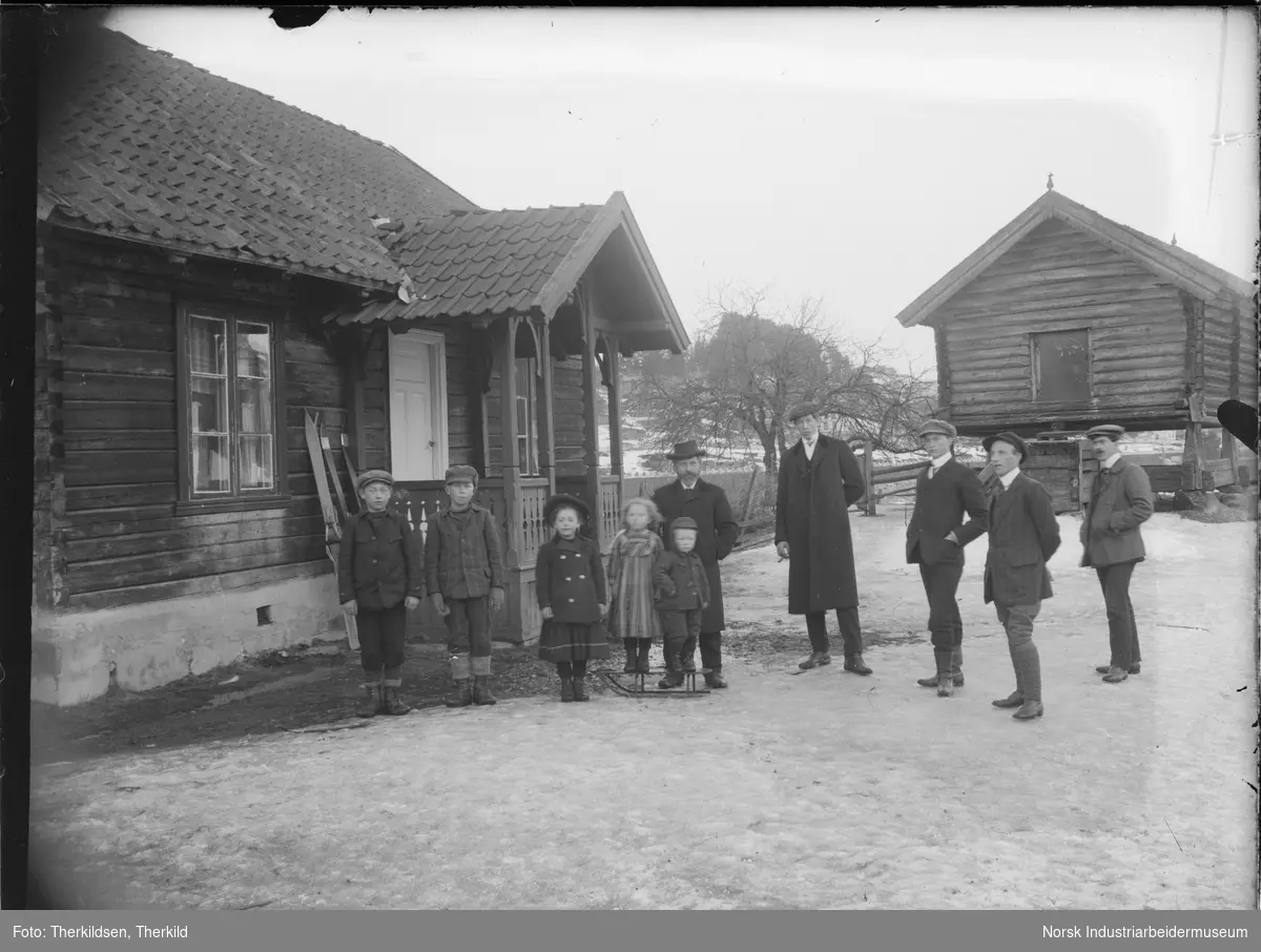 Ti mennesker stående på gårdstun på Sagafoss husmannsplass. Fem barn og fem menn står ved inngangen til bolighuset. Ski står opp mot veggen på huset. Stabbur bak mennene.