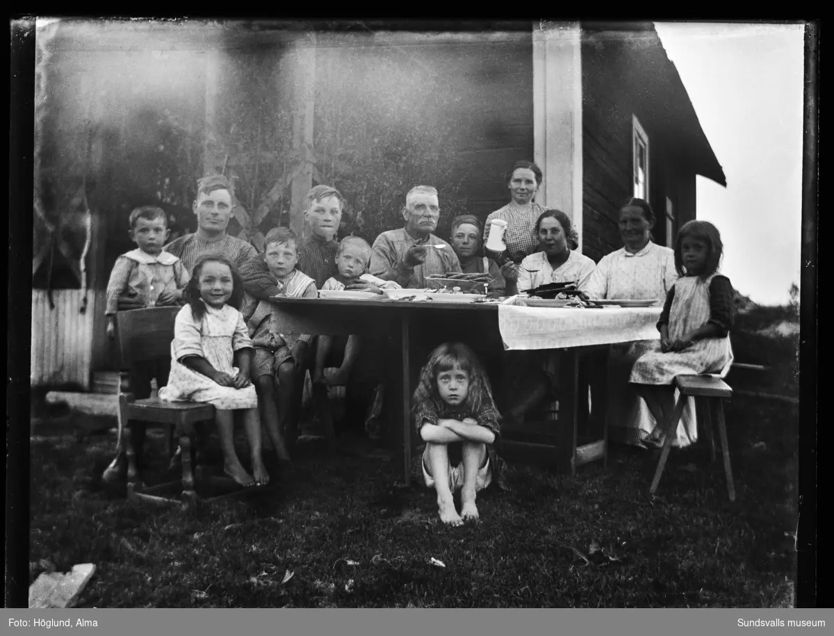 Män, kvinnor och barn vid ett bord intill en timmerstuga. Troligen är det äldre paret fotografens svärföräldrar Per Höglund med hustru. Längst fram sitter tvillingdöttrarna Ingrid och Irma Höglund.