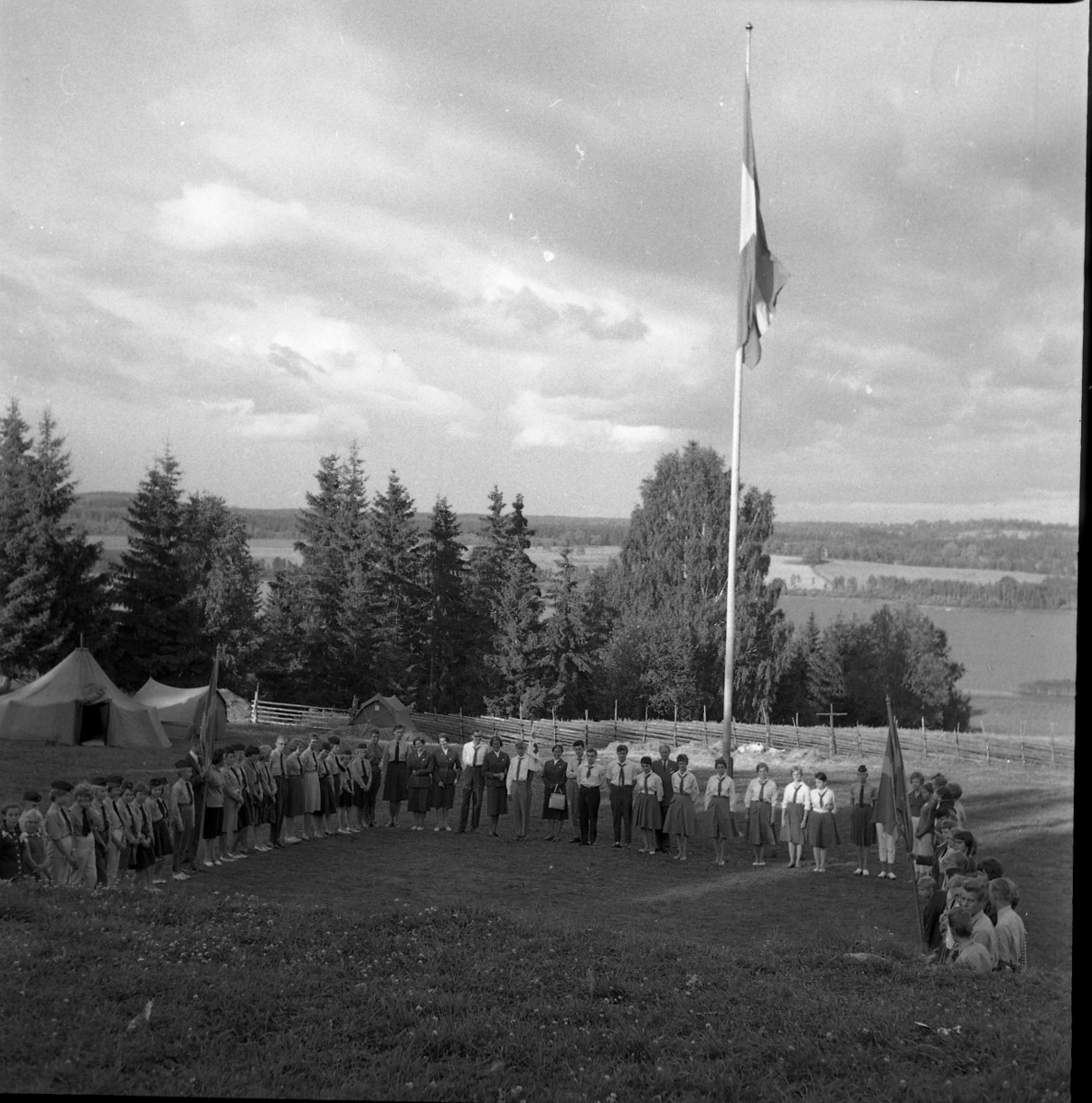 Vy mot en sjö. I mitten står en flaggstång och runt den står det kvinnor och män. Vid sidan av den långa raden med människoir står ett tält upprest.