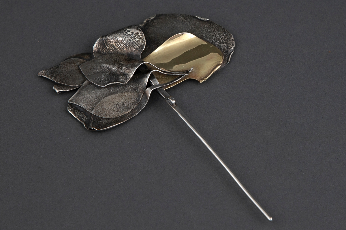 Brosje med stiliserte bladformer av smidd sølv, samt en gullplate, festet til en lang nål. Sølvet har en ru, matt overflate, mens gullet er blankt.