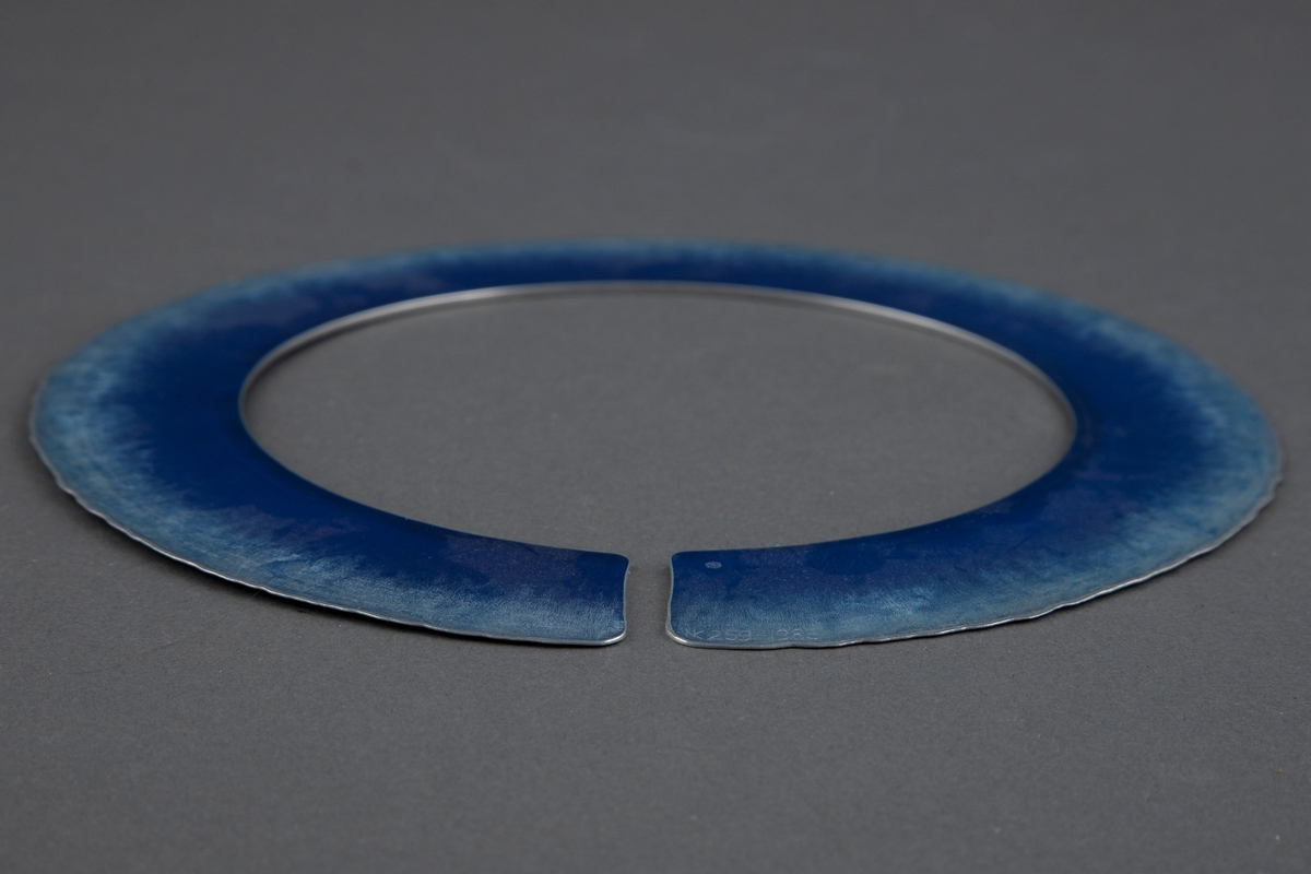 Halssmykke bestående av en cirka 3 cm bred aluminiumsplate i sirkelform med en liten åpning. Begge sider av platen har en dyp mellomblå farge innerst. Fargen blir gradvis lysere mot yttersiden, og den ender i en svakt, bølgeformet kant ytterst.
