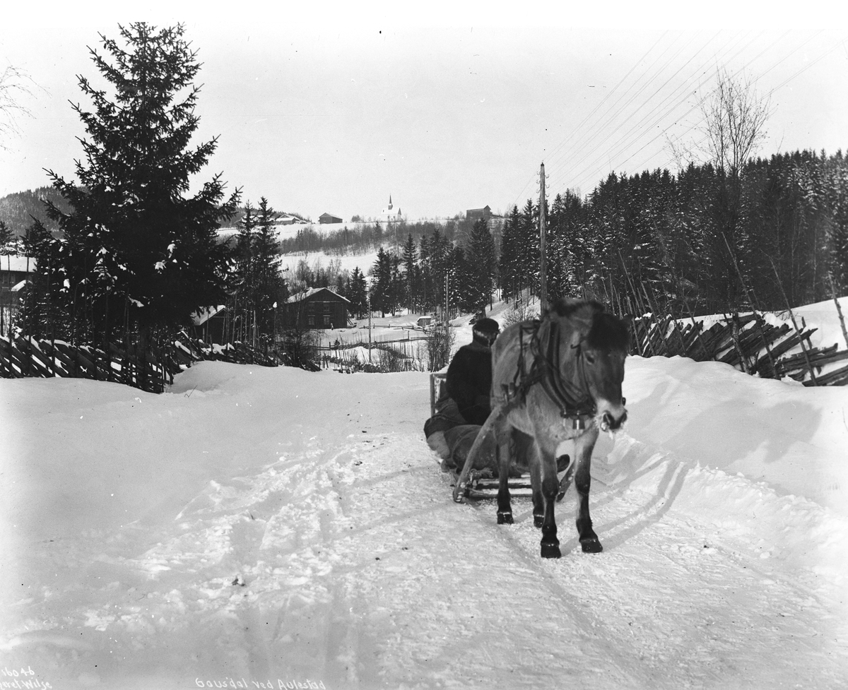 Godt kledd hestekjører i slede på veien fra Aulestad gård med Follebu kirke oppe på høyden i bakgrunnen, Gausdal, Oppland, 1913.