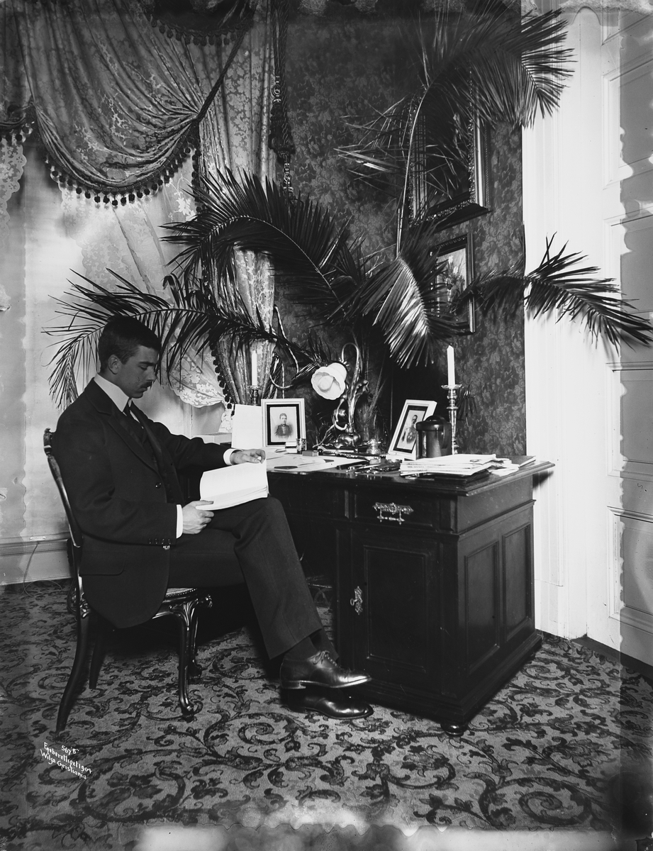 Prins Gustav Adolf ved sitt skrivebord, grønne palmer og omfangsrike portierer i henhold til tidens mote.
