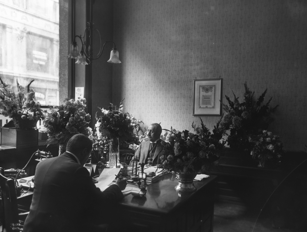 Kontoret bærer preg av festligheter og jubileum, de to herrene sitter omringet av blomsterdekorasjoner.