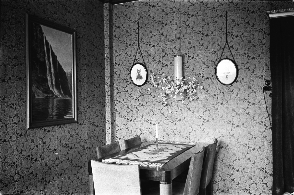 Del av stue, spisebord med vevet løper og fem stoler rundt bordet.  På veggen to bilder og lampett i midten, glimt av døråpning, maleri på den andre veggen.