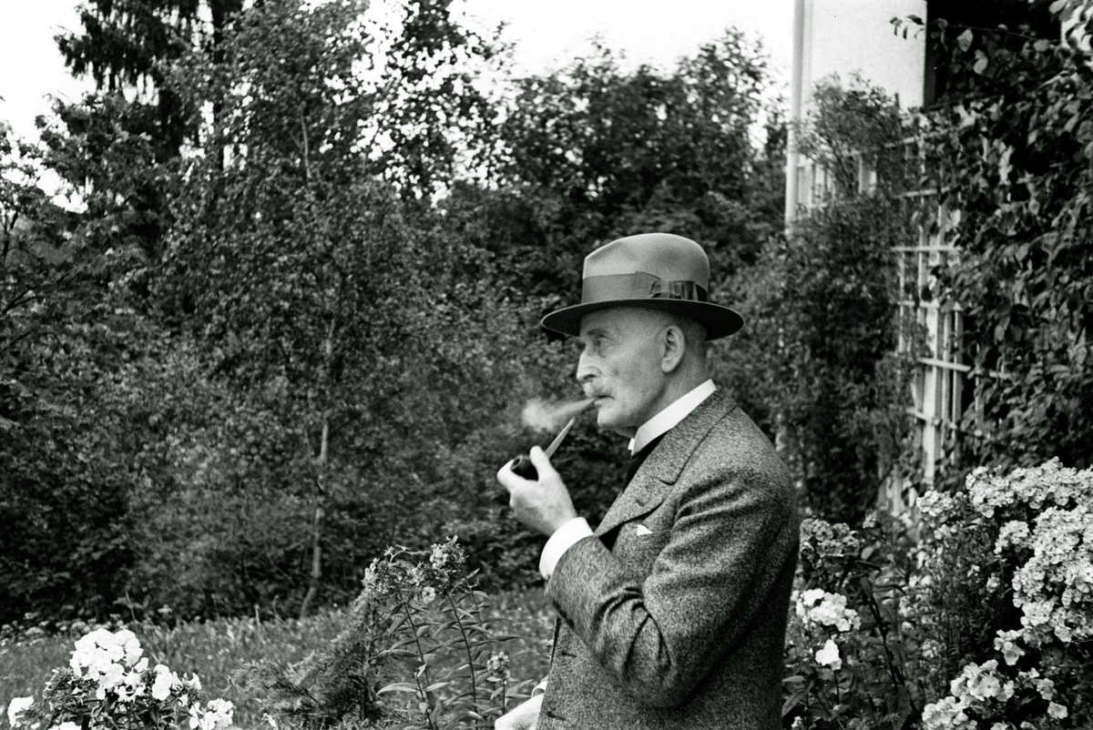 Knut Hamsun nyter sin pipe i husveggen.  Tidsmessig antrukket i dress og hatt. Hamsun hadde en vakker bolig i sjøkanten mellom Lillesand og Grimstad.