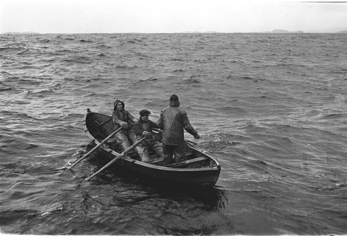 3 mann i sjekte prøver fiskelykken. To mann ved årene og en prøver dorgen bakerst i båten.
