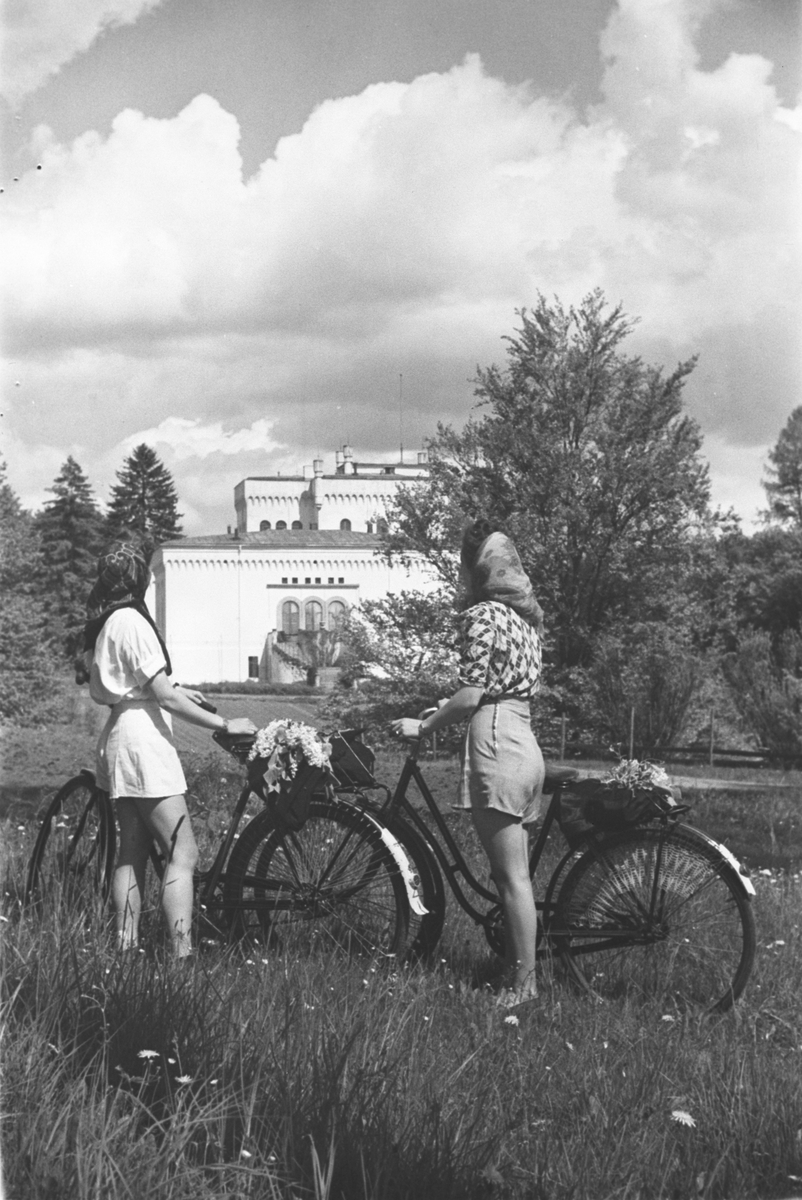 To kvinner på sykkeltur i området ved Bærum Verk.  I bakgrunnen ser vi  hovedgården som tilhører  jernverket.