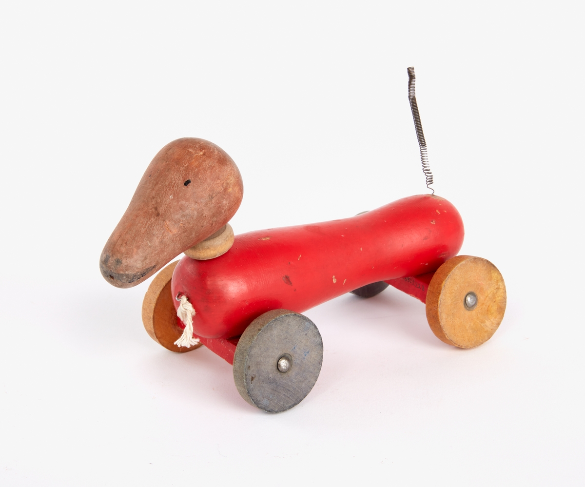 En hund av trä. Långsmal rödmålad kropp med omålat huvud och fyra hjul. Svans av metallfjäder.