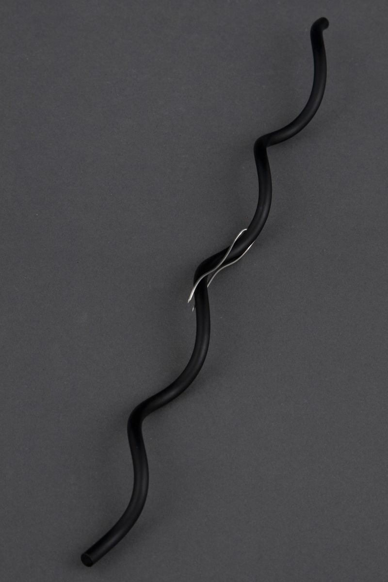 Brosje i form av en bølget, svart aluminiumsstav. Gjennom denne er det stukket en dobbel, tynn ståltråd som nål for å feste brosjen.