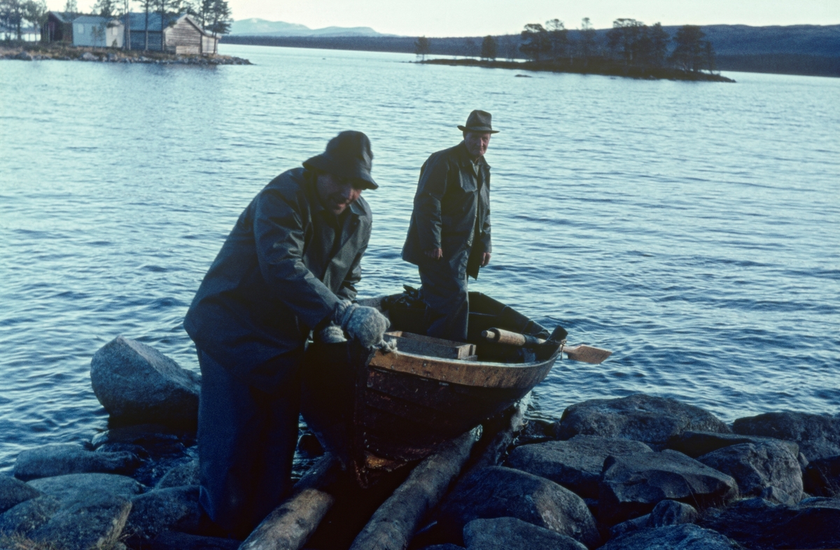 Båt ved land, fra Fiskevollen ved Sølensjøen i Øvre Rendalen i Hedmark. Haagen Hangaard (1899-1989), fotografert stående i en av båtene sine mens den ble trukket opp på den steinete stranda ved Fiskevollen i den nordvestre delen av sjøen. Hangaard eide Enkjegarden eller Nord-Berge i hovedbygda tre mil lengre vest, og var dermed blant dem som hadde fiskerett i denne innsjøen. I tiåra etter 2. verdenskrig ble han ofte omtalt som «Sølensjø-kongen», fordi han i denne perioden var den mest aktive av lotteierne i fisket. Da fotografiet ble tatt var Haagen Hangaard kledd i regnjakke, og han hadde hatt på hodet.