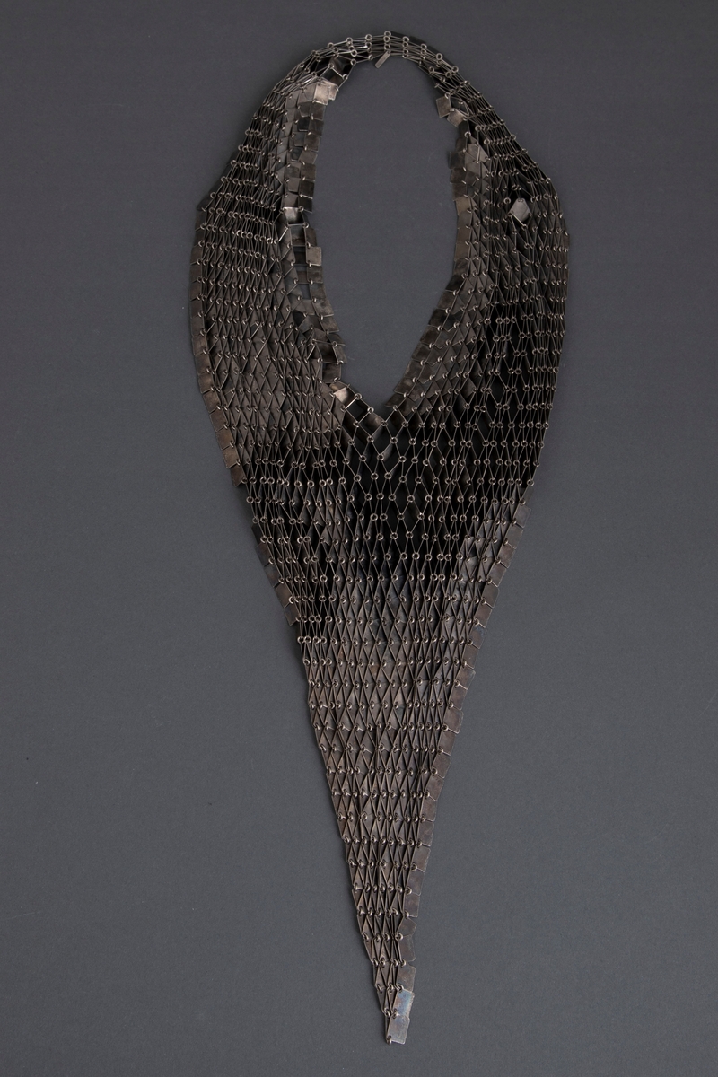 Halssmykke bestående av små rektangler (1 x 0,8cm) i oksydert sølv. Rektanglene er festet til hverandre med øskener. Smykket er utformet som en krage i V-form og ender i en spiss foran. Smykket trees over hodet.