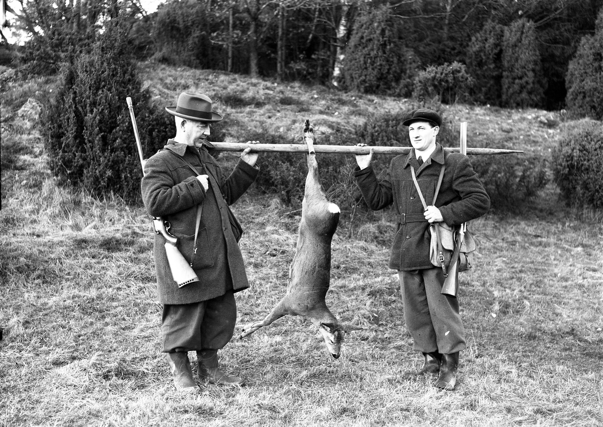 Två jägare, Anton Antonsson och Rolf Halltorp, står i en skogsbacke med nedlagt byte; ett rådjur som hänger på en stör mellan männen.
