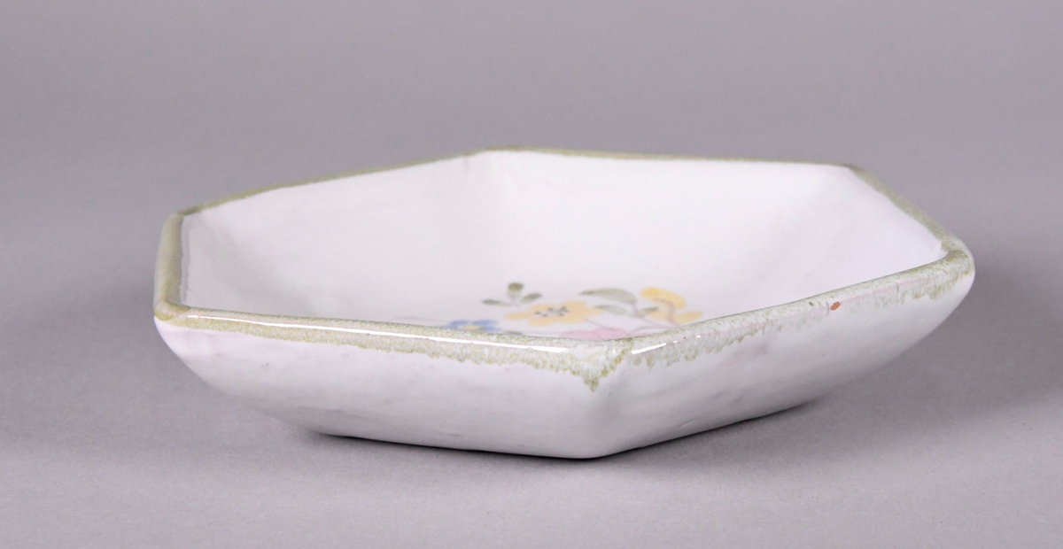 Skål av glassert keramikk, med opphøyd rand. Skålen er sekskantet, og har hvit grunnfarge. Håndmalt dekor av gule, røde og blå blomster, og en grønn borde langs randen.
