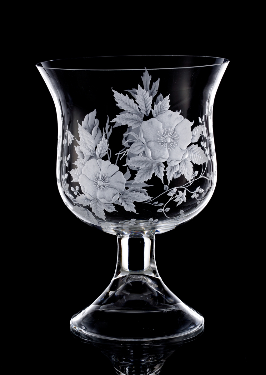 Formgiven av Sigurd Persson, gravyr av Lisa Bauer. Tunn klockformad blomstervas på fot. Gravyrmotiv av linnéa och två andra blommor på kvist.
