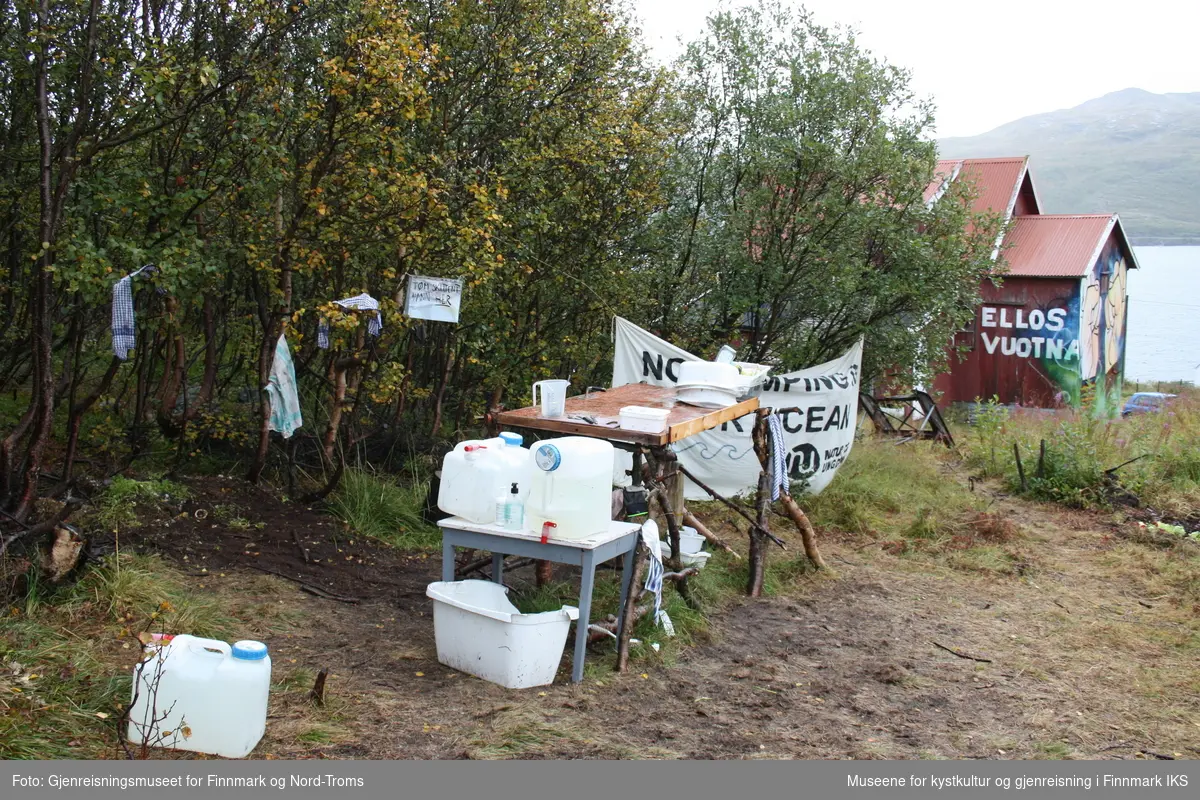 Protestleiren på Markoppneset i Finnmark i 2021. Protestbevegelsen mot dumping av gruveavfall i Repparfjorden har samlet seg og har etablert en teltleir. Bildet er del av en serie som dokumenterer leiren og omgivelsen i området.
Bildet på naustveggen er et verk av kunstneren Tegson og har tittelen "Ellos vuotna".