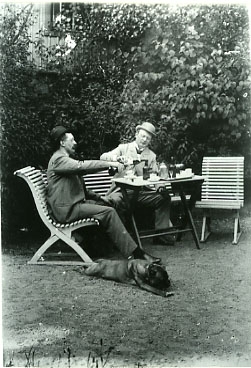 August Bondeson och Hjalmar Carlson sitter i en trädgårdsmöbel och slår i glasen ur buteljer. Båda bär kubb. På marken ligger en hund.