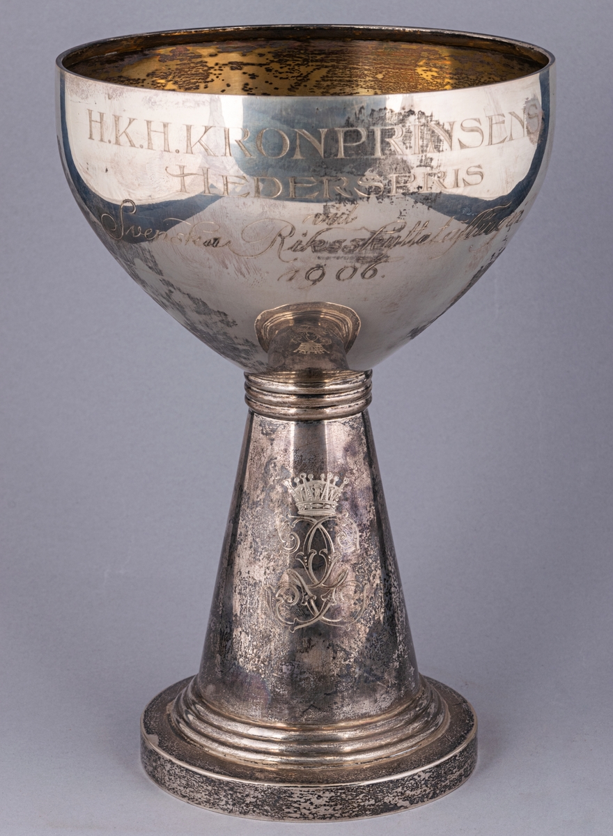 Pokal, silver, inskription: HKH Kronprinsens Hederspris vid Svenska Riksskyttetävlingen 1906. 
På foten kronprinsens krönta spegelmonogram.