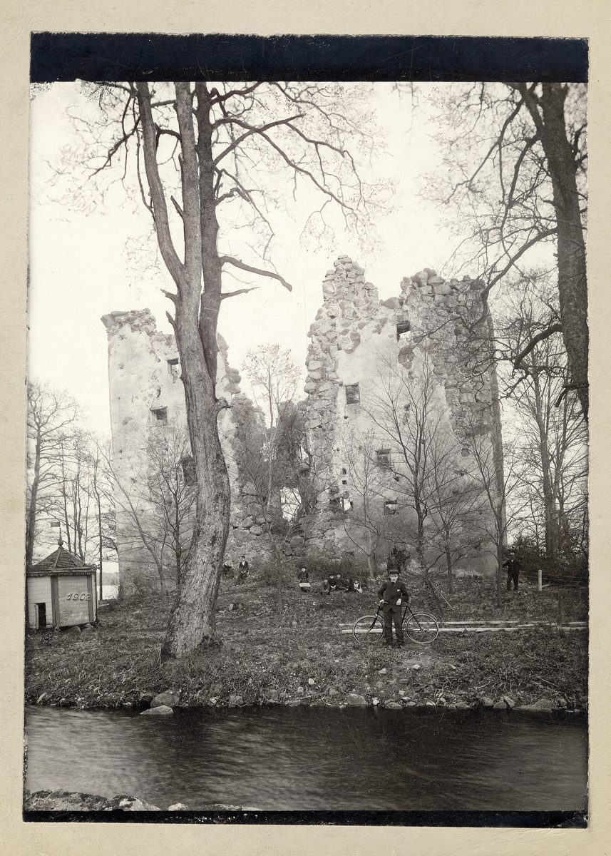 En ung man med cykel poserar vid Bergkvara slottsruin. I bakgrunden syns ytterligare några personer.
I nedre vänstra hörnet är ett årtal instämplat: "1902".