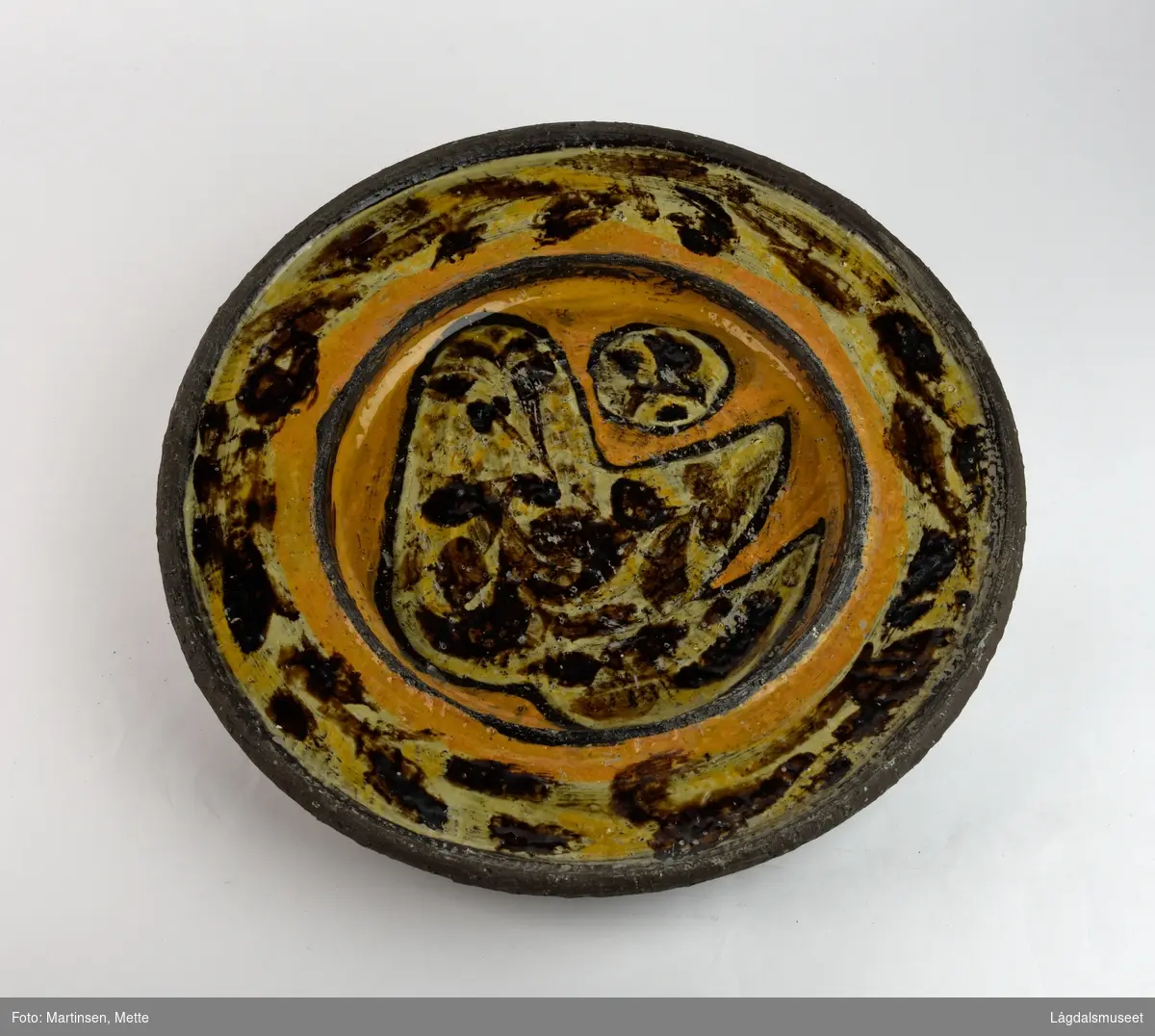 Rundt keramikkfat i brune farger i chamotteleire. Motivet er åpent for tolkning, men ligner på en fugl vendt mot venstre. Over og til venstre for fuglen er en rund figur, et egg? På baksiden hull og metalltråd for oppheng.