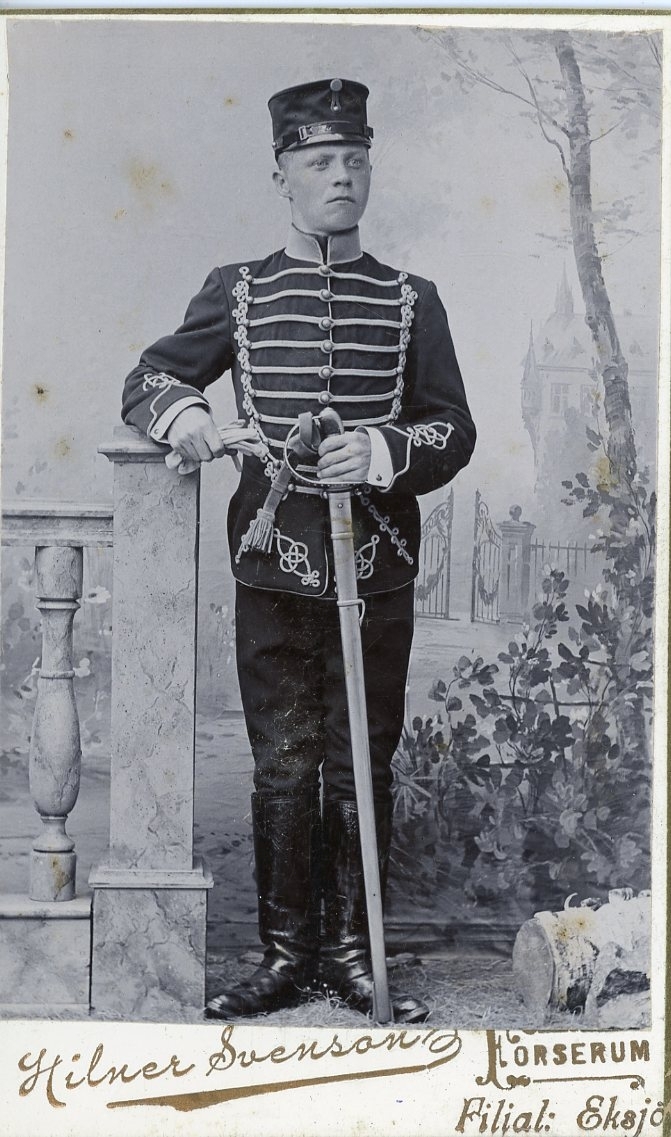 Kabinettsfotografi av en ung man i husaruniform, möjligen modell 1895 Skånska husarregementet. Han står med en värja framför sig.