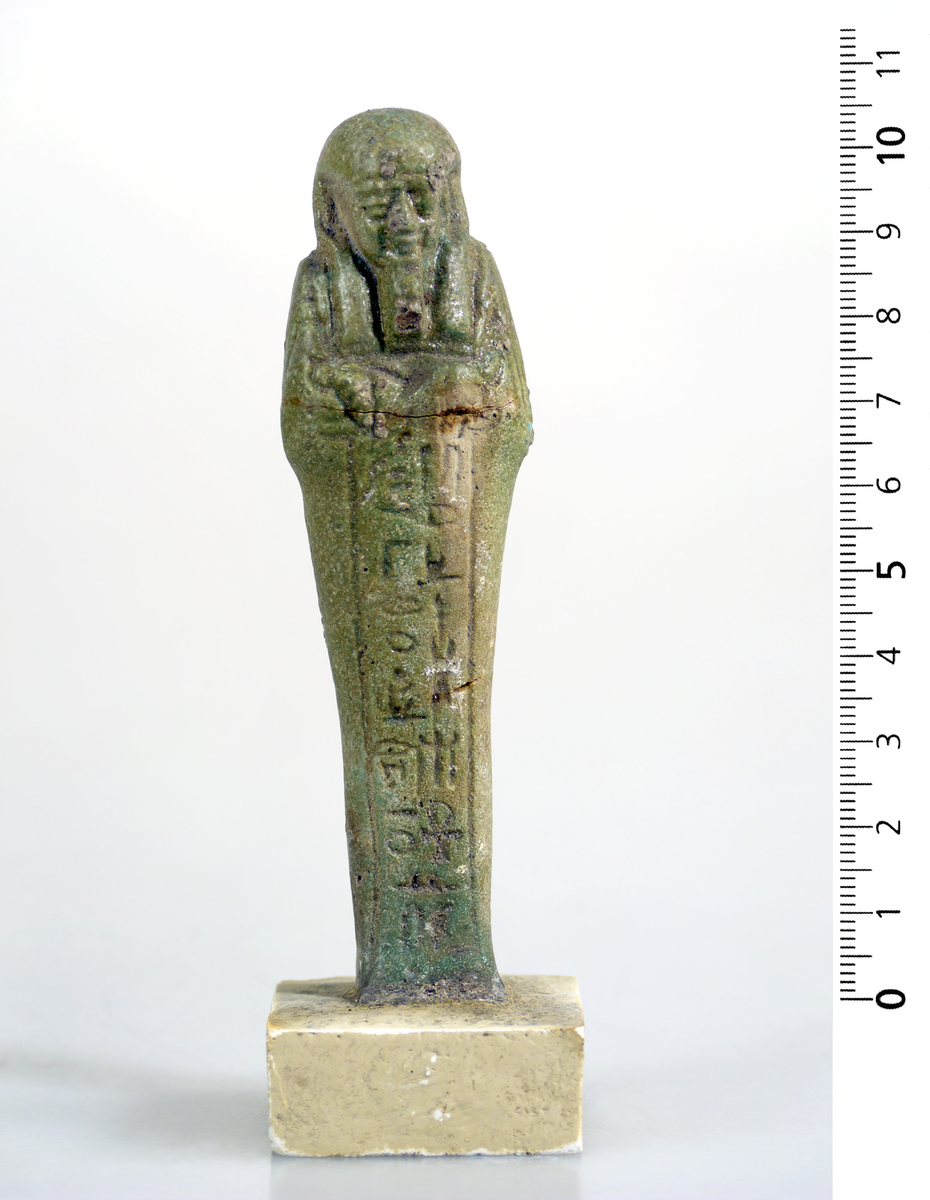 Ushabti i mørk grønn fajanse med hieroglyfer foran. 10,5 cm høy.
Fajanse