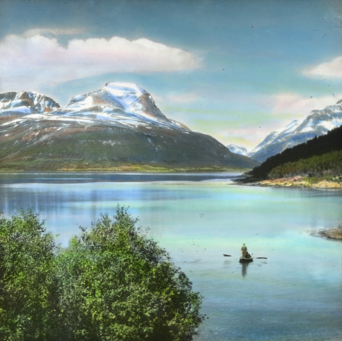Håndkolorert dias. En robåt med to menn på et vann eller en fjord ved Båtsfjord. En av mennene står i båten, mens den andre ror. I bakgrunnen sees snødekte fjell.