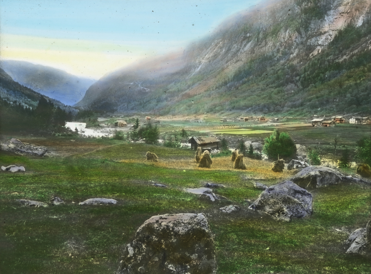 Håndkolorert dias. Åkre og gårder ligger spredt utover dalen mellom bratte fjellsider ved Såheim på Rjukan i 1907.