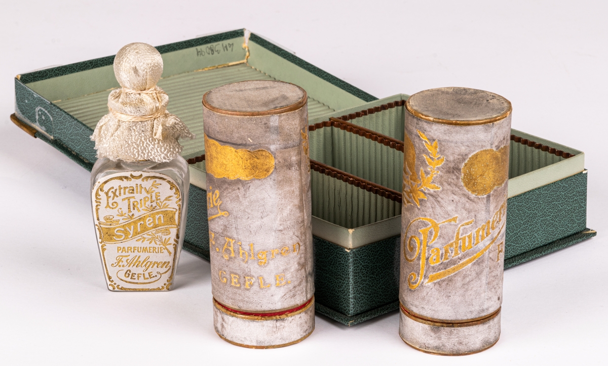 Grön ask innehållande tre parfymförpackningar. Text i guldtryck på asken "Parfumerie F. Ahlgren GEFLE".  En glasflaska och två runda pappförpackningar utan parfymflaskor.