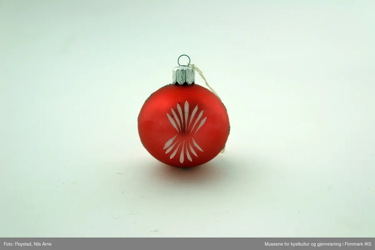 En liten julekule i hard plast med en halvmatt rød/mørkrosa farge med dekor. Dekoren består av påmalte hvite striper på begge sider av kulen og har et blomsterlignende mønster. I tillegg er det dråpeformet dekor malt rød og grønn med glitterkant som går vertikalt rundt kulen, og danner tilsammen en linje. Julekulen har et oppheng av metall med en hvit bomullstråd. Opphenget har gravert mønsterdekor.
