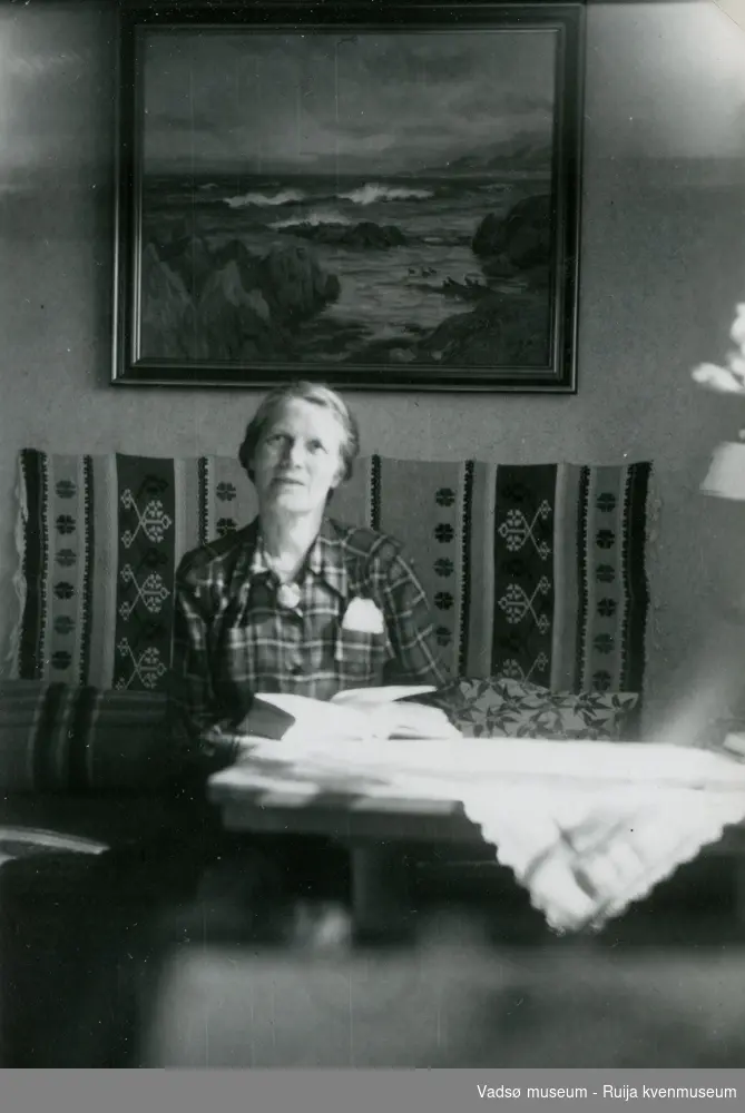 Lilly Simonsen fotografert i sitt hjem i 1953. Lilly sitter på en divan med et vevd teppe bak seg og et Ivar Sælø maleri på veggen bak.