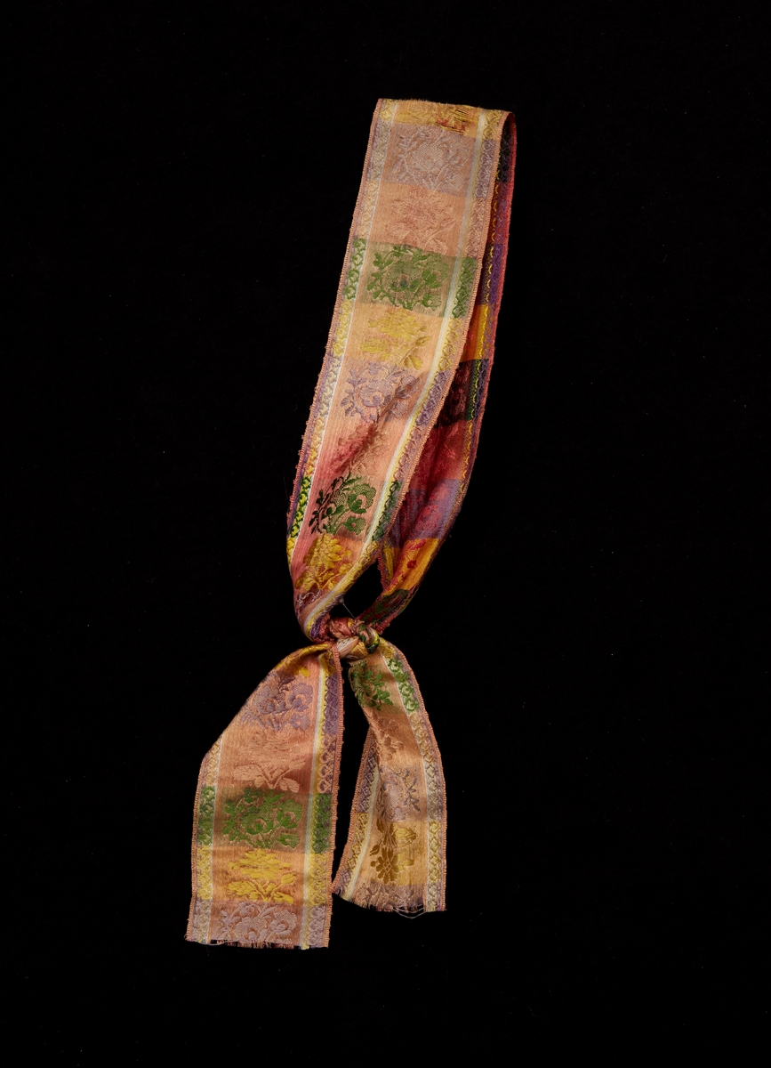Band av mönstervävt siden, tvärrandigt i breda gula, gröna, lila och rosa ränder som har vävda blommönster i varje rand. I kanterna längsgående smala ränder i samma färger som de tvärgående. Knuten ( se bild 1)
Med knut och löst hängande band bäres det runt mössan till Svärdsjö-Envikens kvinnliga sockendräkt (se bild 2 och 3)