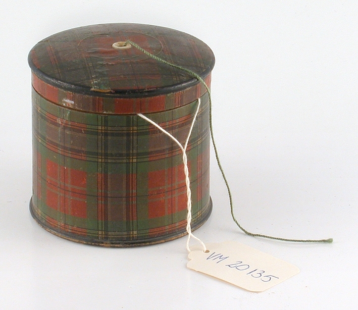Garndosa, överklädd med mönster papper i skotskt mönster med hål i locket för trådens löpande. Avsedd för Clark & Cos ankartråd, firmamärke inuti locket, innehåller två små spetsar.