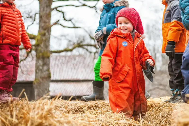 Barn med oransje parkdress og rød nisselue leker i høyet på Follo museum.