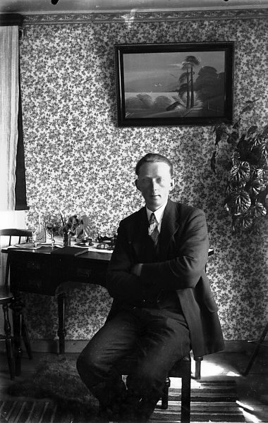 En man sitter vid ett skrivbord i ett rum med småblommig tapet. På väggen hänger en tavla med stiliserat landskap. (Se även bildnr HB504, HB505)