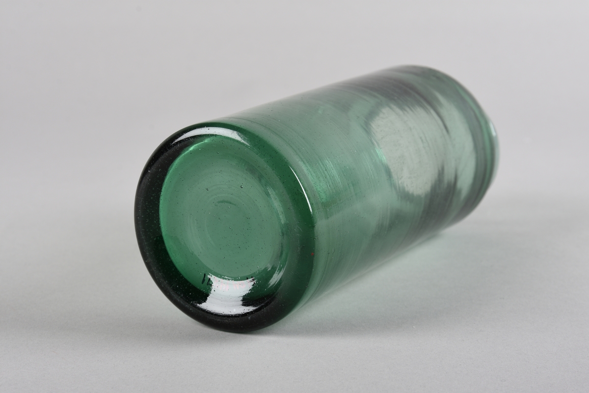 Sylindrisk støypt glaskrukke. Korpus snevrast inn i toppen og deretter ut til ein smal ujamnt forma krage ved munningsranda. Produsert av resirkulert glas. Glaset er grønfarga.