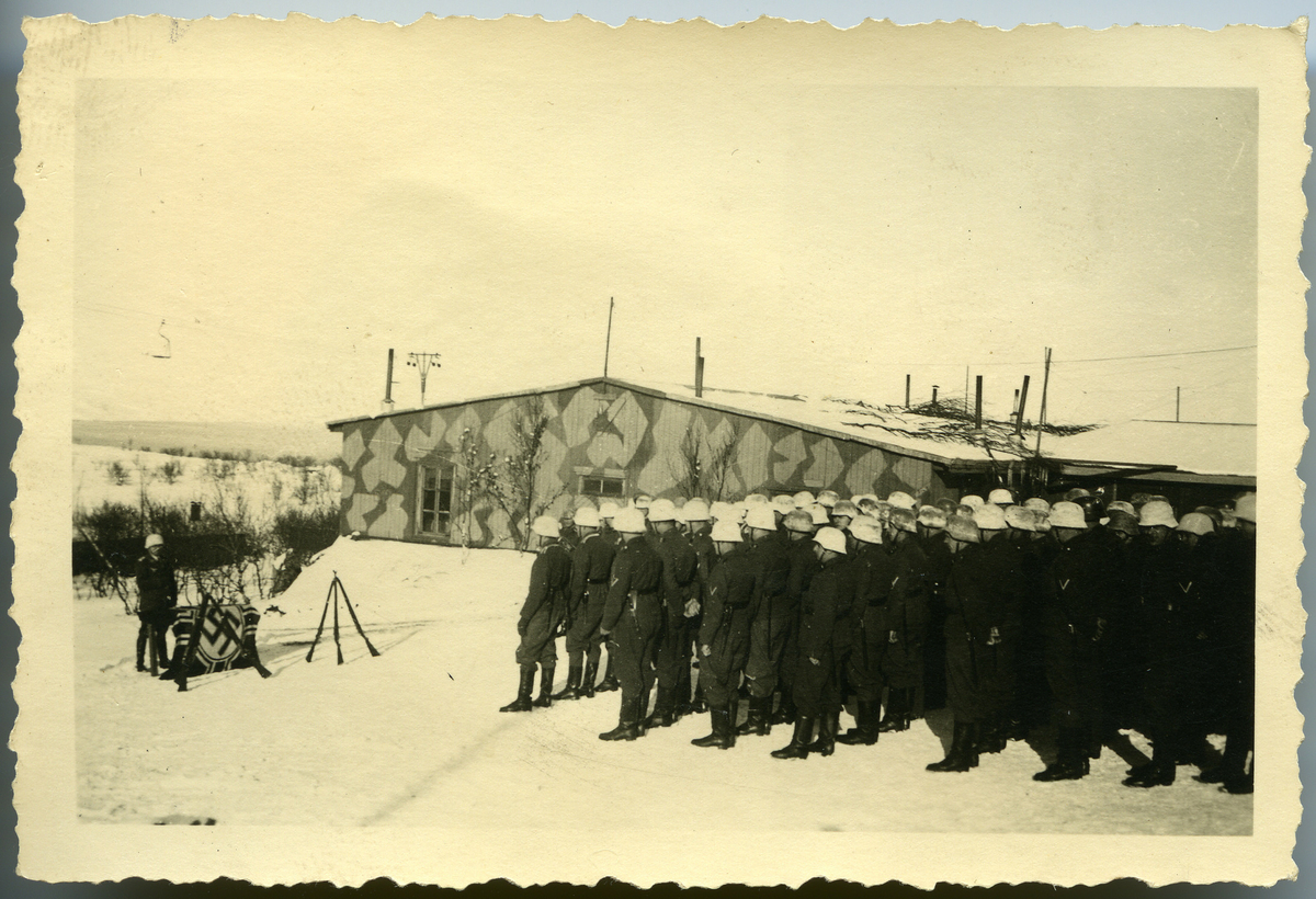 Tyske soldater har tatt oppstilling ved siden av et brakkebygg. Foran dem  står det en soldat bak en hakekorsflagg og gevær som er satt opp tre og tre. Det er vinter. Brakka er mal med et kamuflerende mønster.