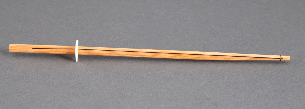 Brosje bestående av en ubehandlet spisepinne i bambus med en halvsirkelformet elfenbensplate plassert litt inn på pinnen fra den tykkeste delen. En svart tråd løper fra spissen over platen og er borret ned i pinnen ikke langt fra den andre spissen. Lang nål på baksiden.