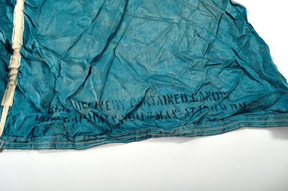 Del av fallskjerm i blå-turkis silke. Trekant forma, sydd saman av fire delar. Merka med 1 2 3 4 ved kvar del. Påsydde kvite band over kvar skjøyt. Kvite snorer