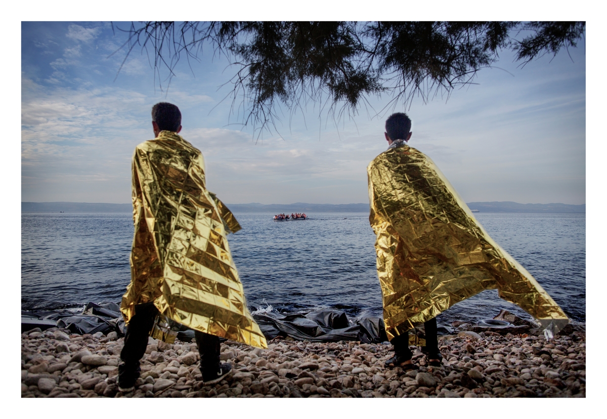 To menn har akkurat ankommet den greske øya Lesvos og har fått utdelt folier for å holde varmen mens de ser to gummibåter med flyktninger nærme seg stranden. Hver dag ankom rundt 2.000 flyktninger, de fleste fra Syria, den lille ferieøya. Turen fra Tyria tar rundt en time i gummibåt og koster flyktningene ca. 1100 dollar. Sommeren 2015 ankom titusenvis av flyktninger og migranter Italia og Hellas og fortsatte reisen mot nord-Europa.