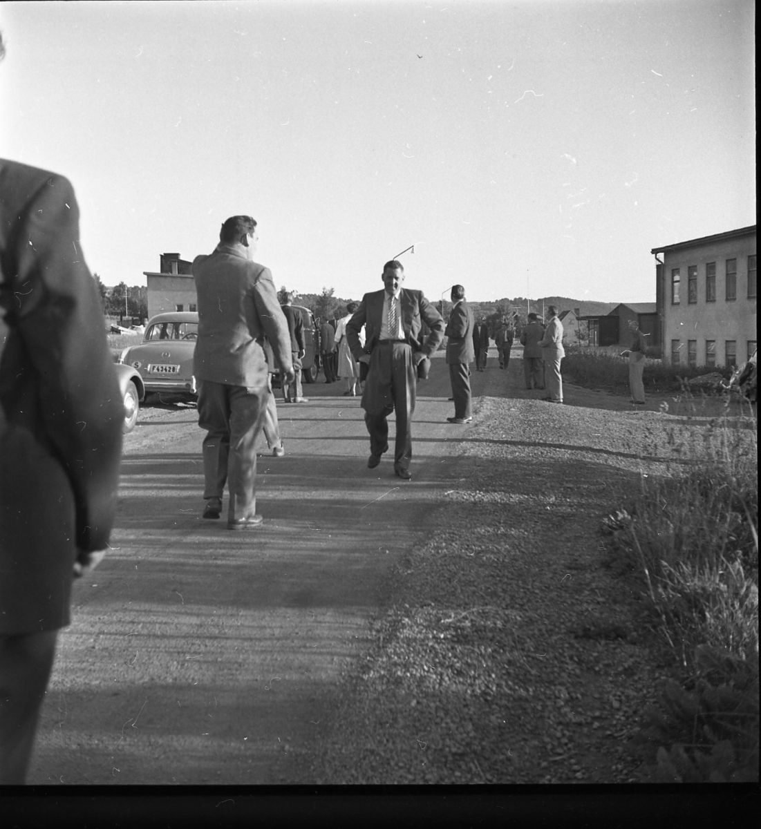Fyra män står på en grusväg. De befinner sig påavstånd från varandra. En bil skymtar i bakgrunden.