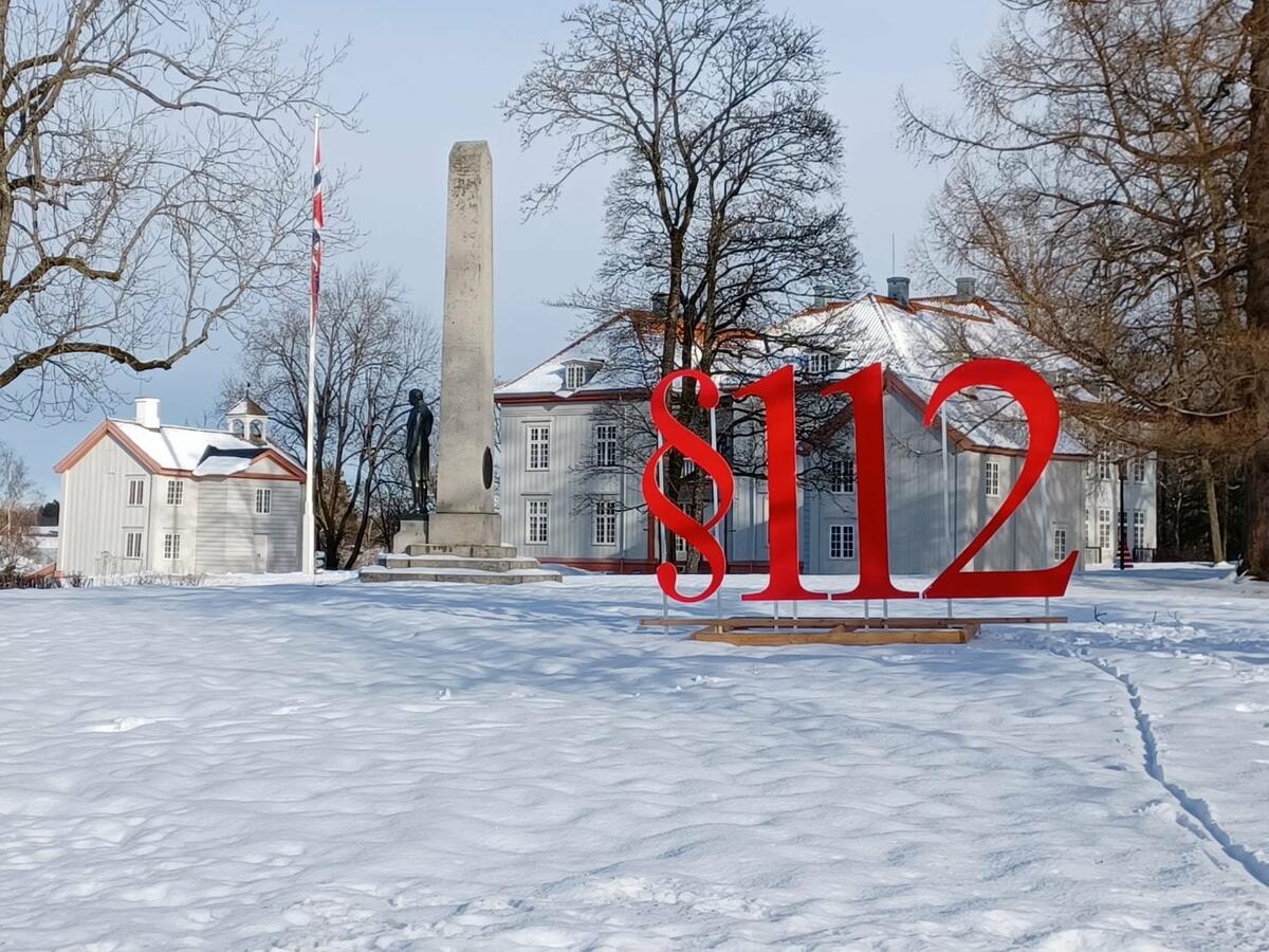 Vinterbilde fra parken med Eidsvollsbygningen i bakgrunnen. Flagg i flaggstanga, sidebygningenr og Carsten Anker-statuen er også i bildet. Snø på bakken, solgløtt. I forgrunnen står §112 i form av to meter høyre bokstaver festet på et stativ.