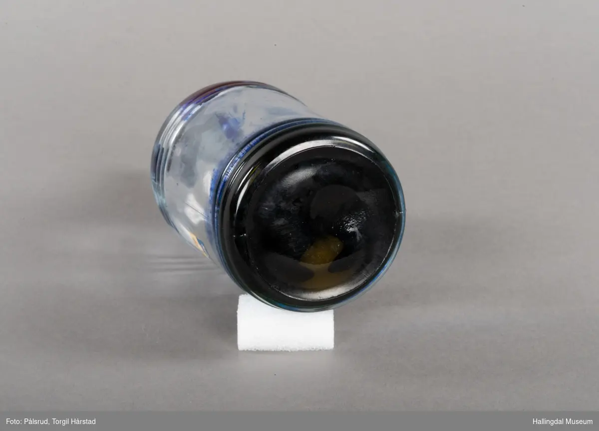 Flaske av blankt glas. Rett, sirkulært tverrsnitt. Det er litt blekk på flaska. Skruvekork av svart plast.