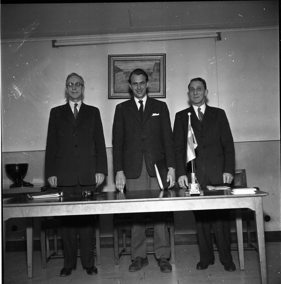 Tre herrar står på rad vid ett bord. På bordet ligger en ordförandeklubba.