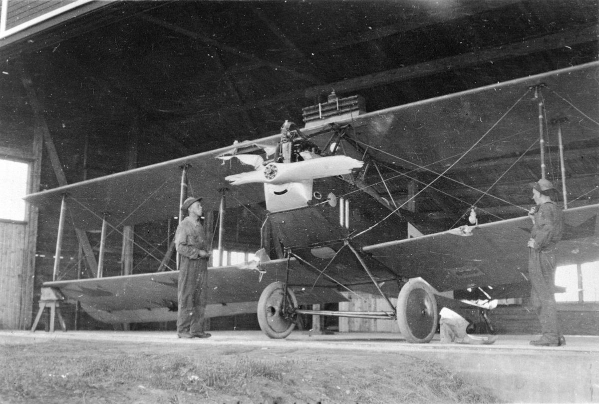 Ett flygplan FVM Albatros 120 med sönderslagen propeller och brustet landningsställ står i en hangar. Efter ett landningshaveri, tidigt 1920-tal. Två flygmekaniker vid planet.