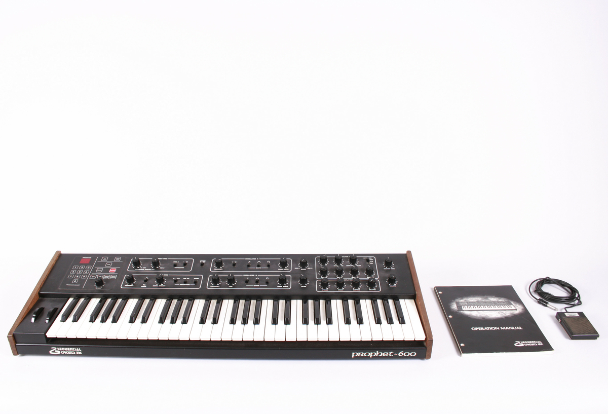 Elektronisk musikkinstrument, synthesizer. Tastatur med klaver nederst mot brukeren. Øverste rekke består av vridbare knapper. På vesntre side et panel med tall. Baksiden har inngang til ulike ledninger.