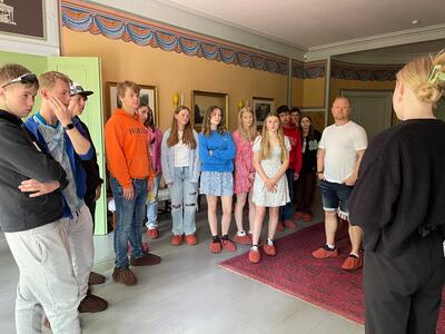 En samling av ungdomsskoleelever fra Vestlandet, iført sommerklær, står i HAgestuen i en ring og lytter til omviseren som forteller.